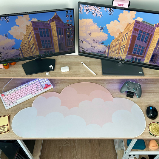Computer setup with dual monitors displaying a cityscape, keyboard, mouse, gaming controller, and peachy beige ombre cloud shaped desk mat on a desk.